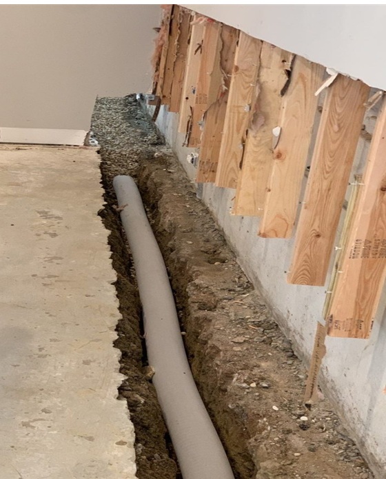 A trench cut into a concrete basement floor containing a new grey PVC drain pipe surrounded by gravel